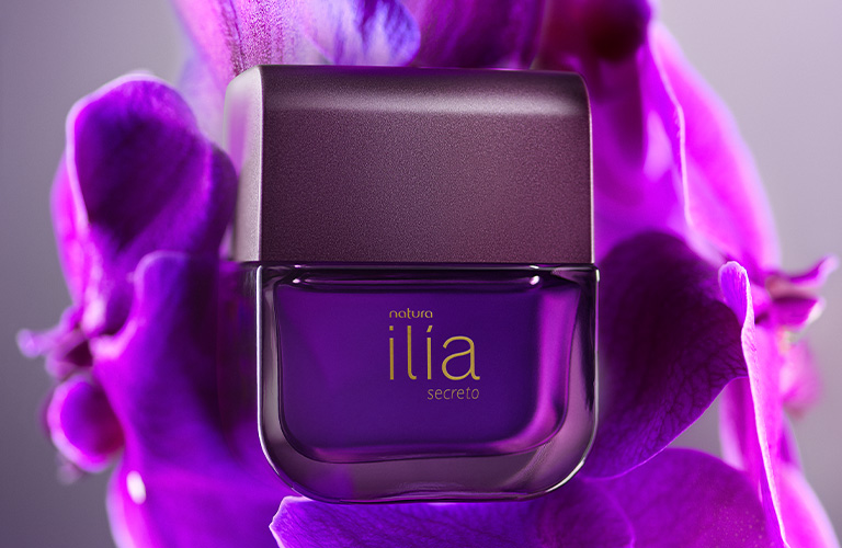 Foto de un frasco de perfume Ilía en color morado al centro. El fondo está compuesto por pétalos de flores en tonos púrpura y lilas, con un fondo difuminado en gris claro.